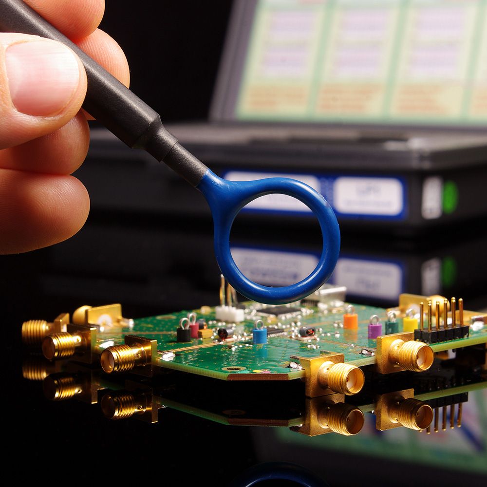 Eine Hand hält eine blaue Lupe über eine grüne Leiterplatte mit goldenen Anschlüssen, die sich auf einer dunklen Oberfläche spiegelt.
